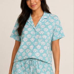 Könnte beinhalten: Ein zweiteiliger Schlafanzug bestehend aus einem kurzärmeligen, geknöpften Hemd mit Reverskragen und einer passenden Shorts. Das Set ist hellblau und mit einem weißen Lotusblütenmuster bedruckt. Die Shorts haben einen Kordelzug am Bund.