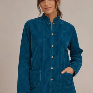 Könnte beinhalten: Eine petrolfarbene Cordjacke mit Stehkragen, langen Ärmeln und einer Knopfleiste auf der Vorderseite mit hellen, runden Knöpfen. Die Jacke verfügt über zwei große aufgesetzte Taschen im unteren Bereich und eine vertikale Rippenstruktur.