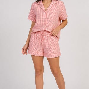 Könnte beinhalten: Ein zweiteiliges Pyjama-Set bestehend aus einem kurzärmeligen Hemd mit Knopfleiste, Reverskragen und Brusttasche sowie einer passenden Shorts mit elastischem Bund. Das gesamte Set besteht aus hellrosa-weißem Vichy-Karo-Stoff.