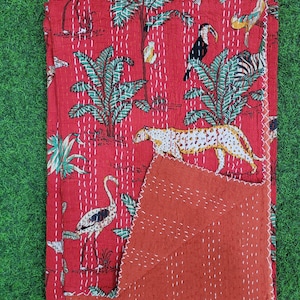 Könnte beinhalten: Eine rote Kantha-Steppdecke aus Baumwolle mit einem Dschungel-Motiv aus Leoparden, Tukanen und tropischen Palmen. Der Stoff ist durchgehend mit weißen Vorstichen bestickt und hat eine einfarbige, rostorangefarbene Rückseite.