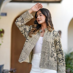 May include: A beige and black patterned jacket with a stand-up collar. The jacket features an intricate floral design and is worn over a white top and white trousers. The sleeves are long and the jacket is open in the front.