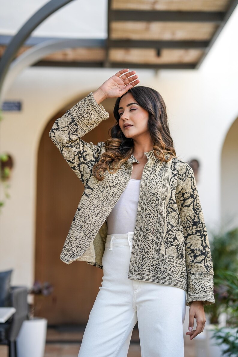 May include: A beige and black patterned jacket with a stand-up collar. The jacket features an intricate floral design and is worn over a white top and white trousers. The sleeves are long and the jacket is open in the front.