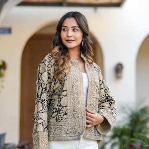 May include: A beige and black patterned jacket with a floral design. The jacket is open, revealing a white top. The model is wearing white trousers. The jacket has a collarless design and long sleeves.
