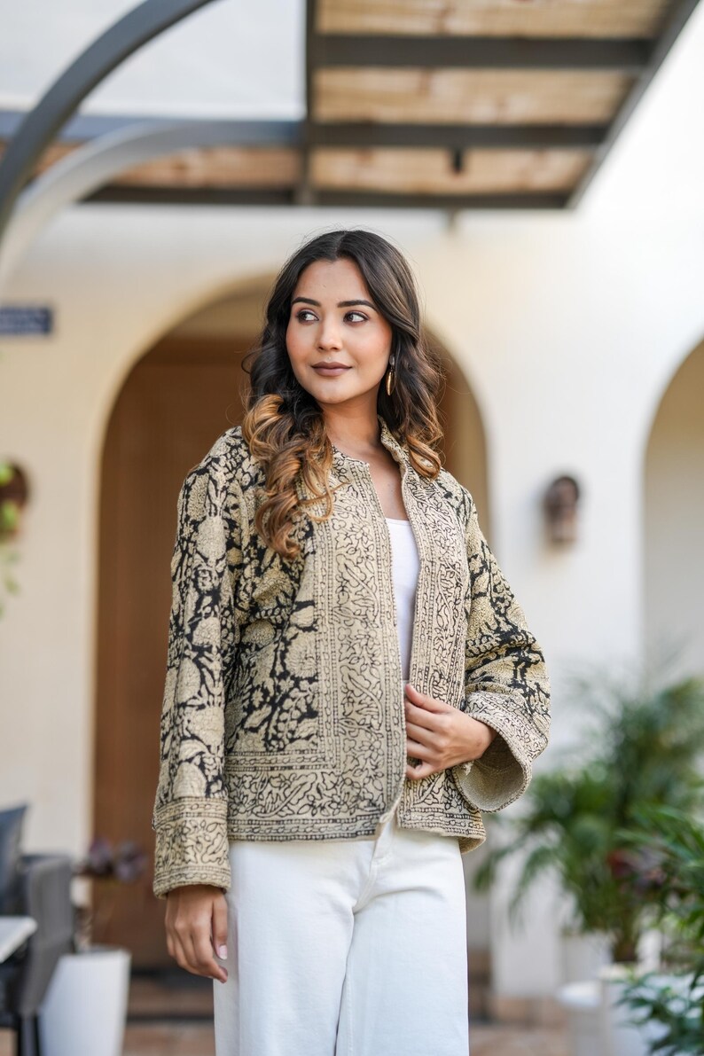 May include: A beige and black patterned jacket with a floral design. The jacket is open, revealing a white top. The model is wearing white trousers. The jacket has a collarless design and long sleeves.