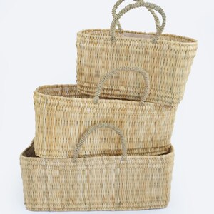 May include: Three woven rectangular baskets stacked on top of each other. The baskets are made of a light tan material with matching handles. The handles are curved and attached to the top of each basket.