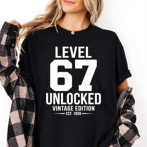 May include: A black short-sleeved t-shirt featuring white distressed-style text that reads LEVEL 67 UNLOCKED VINTAGE EDITION EST. 1959. The shirt is worn with a black and white plaid flannel shirt layered over it, featuring a collar and buttoned cuffs.