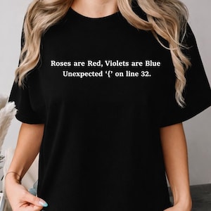 Puede incluir: Una camiseta negra de manga corta con texto blanco impreso en el pecho. El texto dice: Roses are Red, Violets are Blue Unexpected '{' on line 32. La camiseta tiene un cuello redondo y un corte estándar, ofreciendo un aspecto sencillo y limpio.
