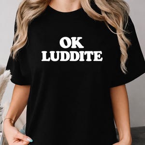 May include: Black t-shirt featuring the white text "OK LUDDITE". The text is in a bold, sans-serif font. The shirt has short sleeves and a crew neck. The text is centered on the front of the shirt.