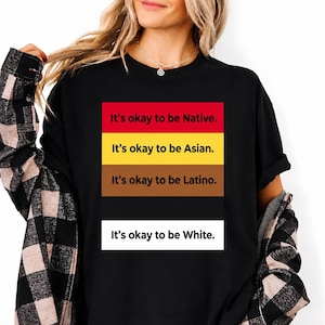 May include: A black t-shirt featuring a graphic with five horizontal color blocks. The text reads: It's okay to be Native, It's okay to be Asian, It's okay to be Latino, and It's okay to be White. The shirt is worn with a black and tan plaid flannel shirt.