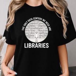 May include: A black short-sleeved t-shirt featuring a circular graphic of a person standing before tall bookshelves. The text reads THE ONLY DATA CENTERS WE NEED ARE LIBRARIES in white capital letters arranged around the central illustration.