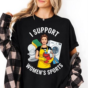 Puede incluir: Camiseta negra con el texto "I SUPPORT WOMEN'S SPORTS" sobre un gráfico de una mujer con un delantal amarillo que sostiene productos de limpieza. Una lavadora, una esponja y platos también están en el gráfico.