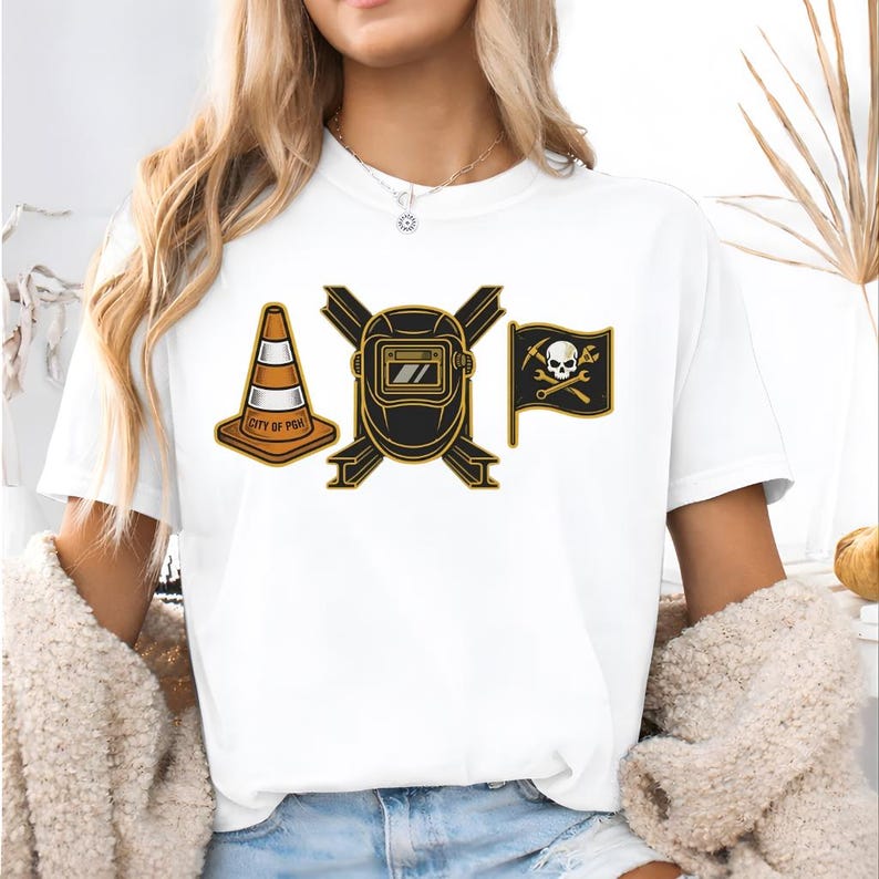May include: White t-shirt featuring three graphic designs: an orange and white traffic cone with "CITY OF PGH" text, a black and gold welding mask, and a black flag with a skull and crossbones. The shirt is made of a soft material.