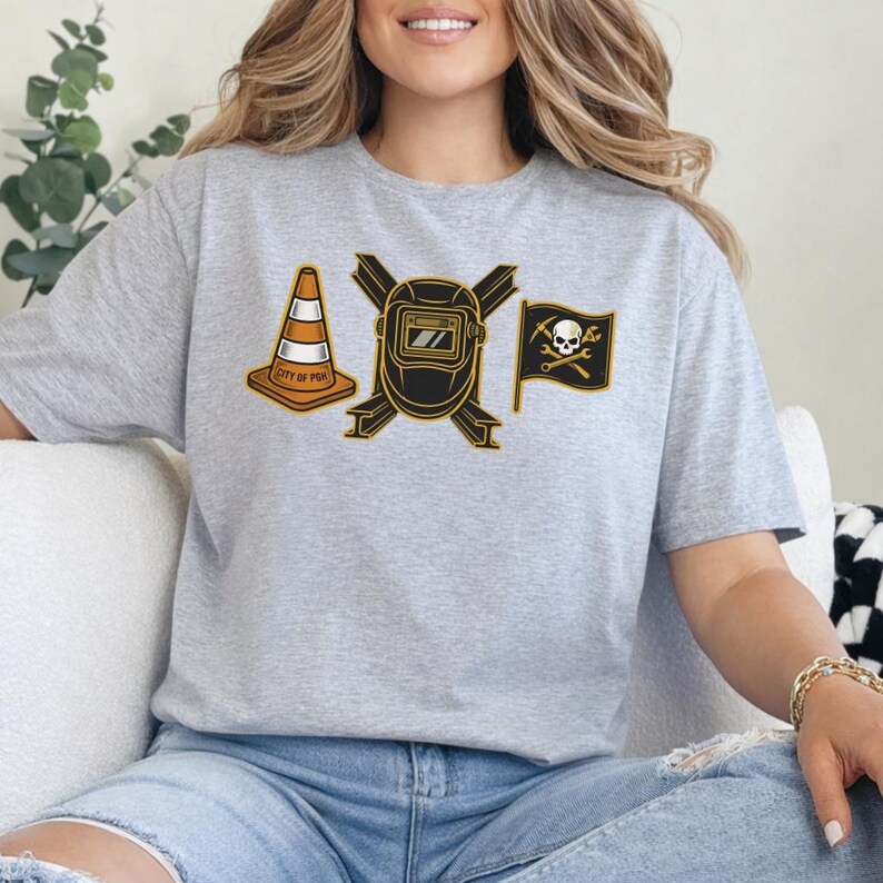 May include: A light gray t-shirt featuring a graphic design with a traffic cone, a welding mask, and a pirate flag with a skull and crossbones. The traffic cone has the text "CITY OF PGH".
