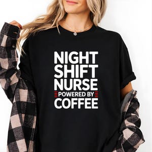 Peut inclure: Un t-shirt noir à manches courtes avec le texte blanc NIGHT SHIFT NURSE POWERED BY COFFEE, accompagné de petites lignes horizontales rouges de chaque côté du mot POWERED. Le t-shirt est porté sous une chemise à carreaux noirs et beiges.