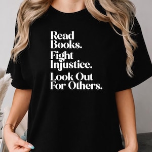 May include: Black t-shirt featuring the white text: "Read Books. Fight Injustice. Look Out For Others." The shirt has a crew neck and is made of a soft fabric. The text is in a bold, sans-serif font, making it easy to read.
