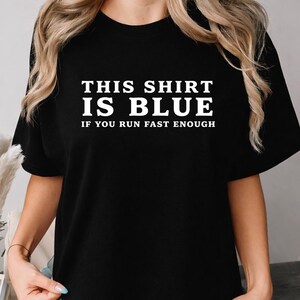 Puede incluir: Una camiseta negra de manga corta con texto blanco impreso en el pecho que dice: THIS SHIRT IS BLUE IF YOU RUN FAST ENOUGH. La prenda tiene un cuello redondo y un corte estándar, mostrando el texto claramente en una fuente sans-serif.