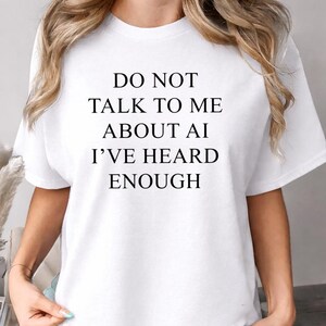 Może przedstawiać: Biały t-shirt z krótkim rękawem z czarnym napisem wyśrodkowanym na klatce piersiowej o treści: DO NOT TALK TO ME ABOUT AI I'VE HEARD ENOUGH. Odzież posiada klasyczny okrągły dekolt i standardowy krój, wykonana z gładkiej białej tkaniny.