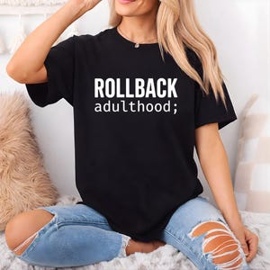 以下が含まれることがあります： 白文字で「ROLLBACK adulthood;」と書かれた黒いTシャツ。カジュアルなスタイルで、普段使いに適しています。テキストは太字のサンセリフ体です。