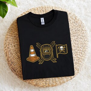 May include: Black t-shirt featuring a gold outline design of a traffic cone with "CITY OF PGH" text, a welding mask, and a pirate flag with a skull and crossbones. The shirt is displayed on a woven surface.