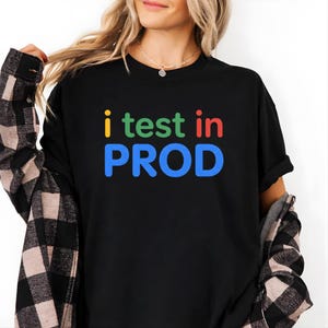 Op de afbeelding: Zwart T-shirt met de tekst "i test in PROD" in geel, groen, rood en blauw. Het shirt wordt gedragen met een zwart-wit geruit overhemd.
