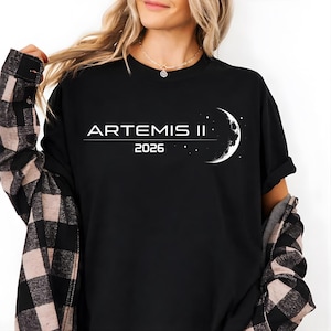 Puede incluir: Camiseta negra con el texto blanco "ARTEMIS II 2026" y un gráfico de media luna. La camiseta es de cuello redondo.