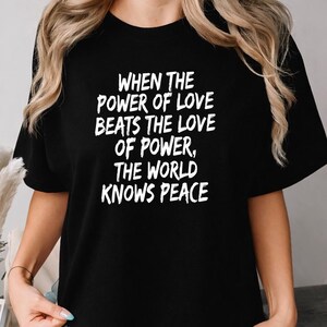 Może przedstawiać: Czarny t-shirt z krótkim rękawem z białym nadrukiem na przodzie o treści: WHEN THE POWER OF LOVE BEATS THE LOVE OF POWER, THE WORLD KNOWS PEACE. Koszulka posiada okrągły dekolt oraz standardowy krój, na którym wyraźnie widać ten cytat.