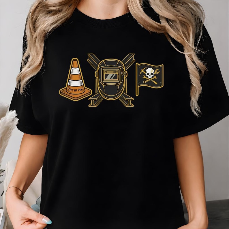 May include: Black t-shirt featuring three graphic designs: a traffic cone with "CITY OF PGH" text, a welding mask, and a pirate flag with a skull and crossed tools. The designs are in shades of yellow and orange.