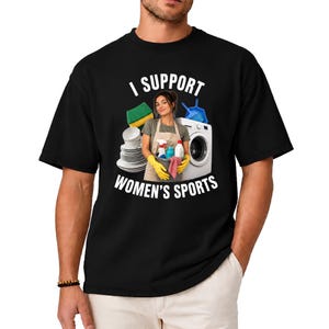 May include: A black t-shirt featuring a graphic of a person wearing an apron and yellow gloves, holding a bucket with cleaning supplies. Beside them are stacks of white plates, a green sponge, a blue dustpan, and a white washing machine. The text reads I SUPPORT WOMEN'S SPORTS.