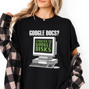 Può includere: T-shirt nero con la scritta "GOOGLE DOCS?" sopra una grafica di computer retrò. Lo schermo del computer mostra "I PREFER TO GOOGLE DISKS" in testo verde. La maglietta è in stile casual.