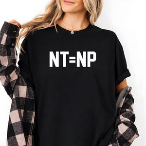 以下が含まれることがあります： 白文字で「NT=NP」と書かれた黒いTシャツ。クルーネックのスタイルです。その上に黒とピンクのチェック柄のフランネルシャツを着ています。