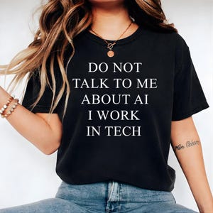 May include: Black crew neck t-shirt featuring the white text: "DO NOT TALK TO ME ABOUT AI I WORK IN TECH." The shirt is made of a soft material. The model is wearing blue jeans.