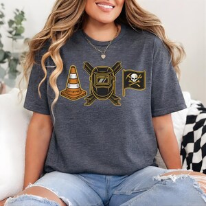 May include: Dark gray t-shirt featuring a graphic design with a traffic cone, a welding mask, and a pirate flag with a skull and crossbones. The traffic cone has the text "CITY OF PGH". The design is in black and gold.