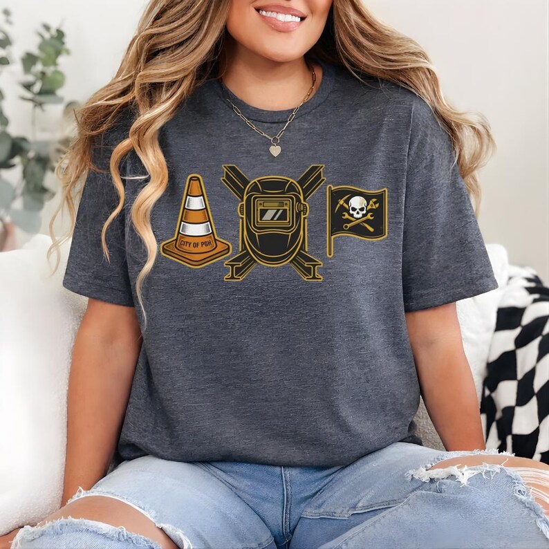 May include: Dark gray t-shirt featuring a graphic design with a traffic cone, a welding mask, and a pirate flag with a skull and crossbones. The traffic cone has the text "CITY OF PGH". The design is in black and gold.