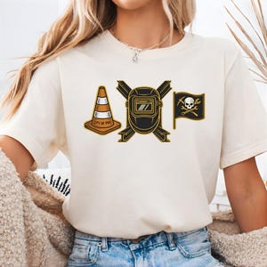 May include: Cream-colored t-shirt featuring three graphic designs: an orange and white traffic cone with "CITY OF PGH" text, a black and gold welding mask, and a black flag with a skull and crossbones. The shirt is made of a soft material.
