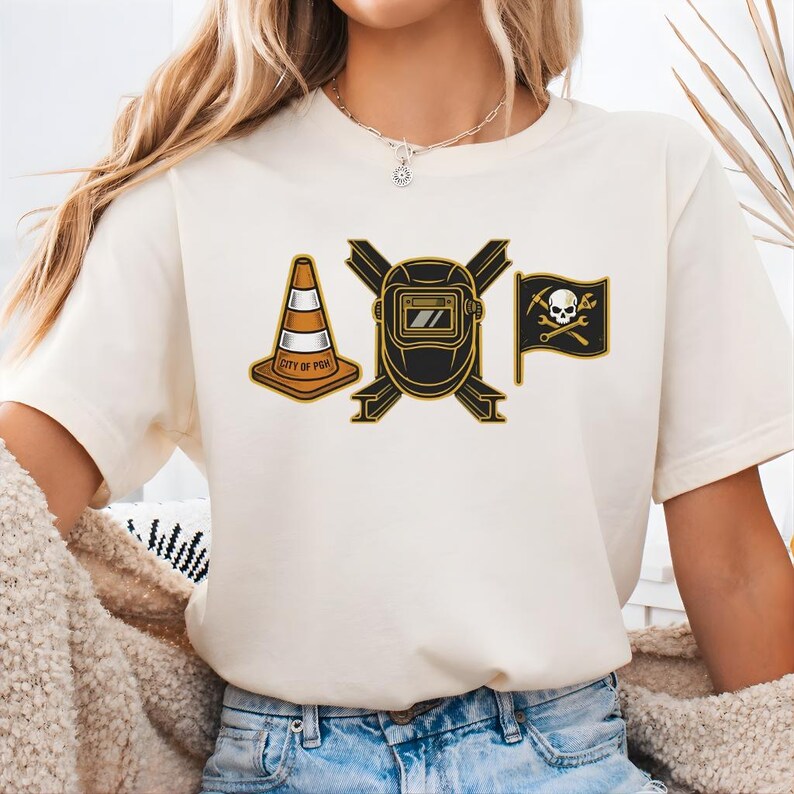 May include: Cream-colored t-shirt featuring three graphic designs: an orange and white traffic cone with "CITY OF PGH" text, a black and gold welding mask, and a black flag with a skull and crossbones. The shirt is made of a soft material.