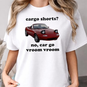 Puede incluir: Camiseta blanca con un gráfico de un coche descapotable rojo y el texto "cargo shorts? no, car go vroom vroom" en negro. El coche tiene faros negros y ruedas plateadas.