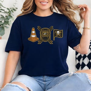 May include: Navy blue t-shirt featuring a gold graphic of a traffic cone, a welding mask, and a pirate flag with a skull and crossbones. The text "CITY OF POR" is on the traffic cone.
