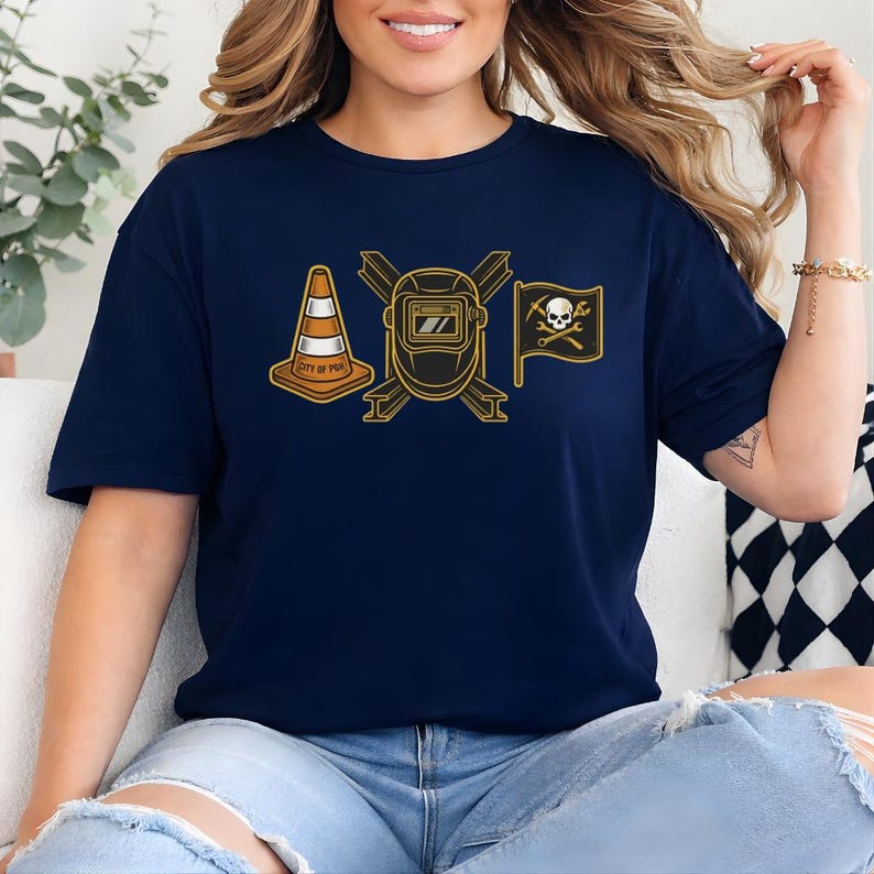 May include: Navy blue t-shirt featuring a gold graphic of a traffic cone, a welding mask, and a pirate flag with a skull and crossbones. The text "CITY OF POR" is on the traffic cone.
