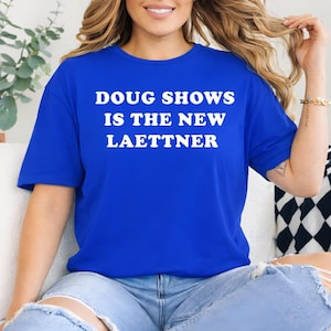 Könnte beinhalten: Ein königsblaues T-Shirt mit dem weißen Text "DOUG SHOWS IS THE NEW LAETTNER". Das Shirt besteht aus einem weichen Material und hat kurze Ärmel. Die Person auf dem Bild trägt eine blaue Jeans.