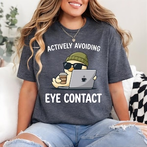 Puede incluir: Camiseta gris oscuro con el texto "ACTIVELY AVOIDING EYE CONTACT". La camiseta presenta un pato de dibujos animados con gafas de sol y gorro, sosteniendo una taza de café y un portátil con un icono de café.