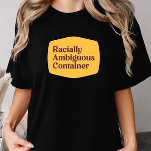 May include: A black short-sleeved t-shirt featuring a yellow, rounded rectangular graphic on the chest. The graphic contains the dark brown text "Racially Ambiguous Container" printed in a clean, sans-serif font centered within the shape.