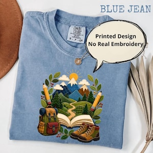 Puede incluir: Una camiseta color azul jean con un diseño estampado de un paisaje montañoso, un libro abierto, una mochila, botas de senderismo y dos lápices. El texto en la camiseta dice BLUE JEAN y Printed Design No Real Embroidery.