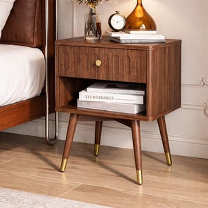 Könnte beinhalten: Ein Nachttisch aus dunklem Holz mit einer Schublade und einem unteren Regal. Die Schublade hat einen goldenen Knopf und ein geschnitztes Design. Der Tisch hat konische Beine mit goldenen Spitzen. Eine Lampe, Bücher und eine Uhr stehen oben.