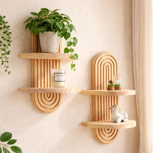 Puede incluir: Dos estanterías de pared de madera con acabado natural. Una estantería contiene una planta en maceta y una vela, la otra pequeños objetos decorativos. Las estanterías tienen un diseño único con detalles arqueados y de listones, ideal para exhibir decoración.
