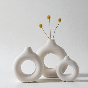 Puede incluir: Tres jarrones de cerámica blanquecina de diferentes tamaños, cada uno con una abertura circular en el centro. Los jarrones están agrupados, con flores amarillas secas en el jarrón más grande. Diseño minimalista y moderno.