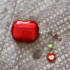 Puede incluir: Una funda roja con forma de manzana con una hoja verde, dise&ntilde;ada para guardar auriculares. Un charm plateado con una peque&ntilde;a manzana, flor, estrella y otros abalorios cuelga de la funda. La funda tiene un acabado brillante.