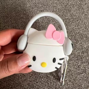May include: White and pink Hello Kitty-themed AirPods case with a bow, headphones, and a yellow nose. The case has a silver clip and a white handle. The case is held by a hand.
