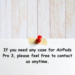 May include: A white note with text about AirPods Pro 3 cases, secured with a red push pin and tan tape, against a light wood grain background.