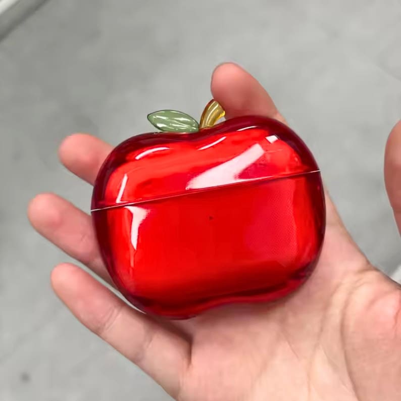 Puede incluir: Un recipiente rojo brillante en forma de manzana con una hoja verde y un tallo amarillo. El recipiente se sostiene en una mano y parece estar hecho de un material brillante. El recipiente es de color rojo vibrante.