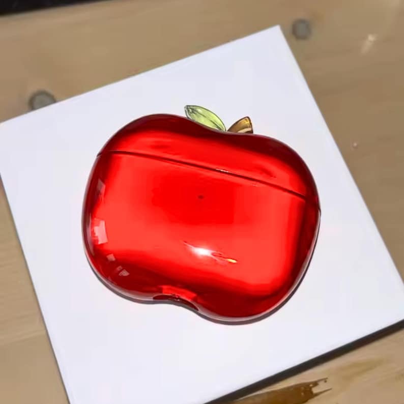 Puede incluir: Una funda roja brillante con forma de manzana con detalles de hojas verdes y doradas. La funda est&aacute; sobre una superficie blanca. La funda tiene una superficie brillante y reflectante.